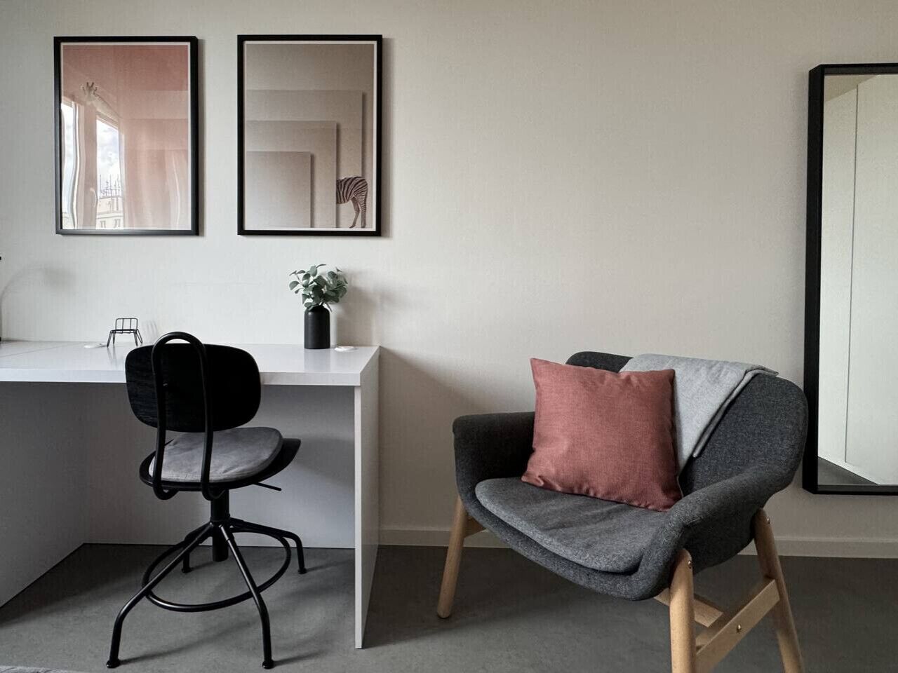 Un espacio de oficina moderno con un escritorio, una silla de oficina negra, un sillón gris con un cojín rosa y dos obras de arte enmarcadas en la pared.