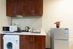 A compact kitchen with wooden cabinets, a microwave, a washing machine, a sink, and a refrigerator. - Mujinhua Hotel (Chongqing)