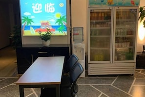 Ein Empfangsbereich mit einer Rezeption, Stühlen, einem großen Bildschirm, der eine Strandansicht zeigt, einem Wasserspender und einem Kühlschrank.