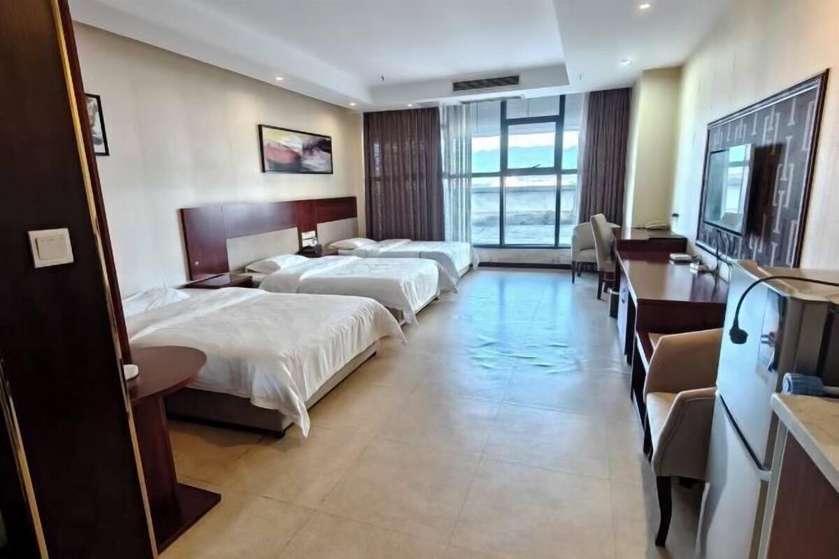 Ein Hotelzimmer mit drei Betten, einem Schreibtisch, einem Fernseher und einem großen Fenster mit Blick.