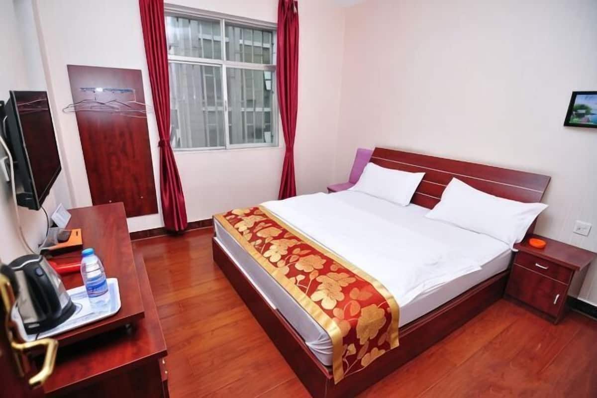A hotel room with a bed, a desk with a kettle, a water bottle, a TV, and a window with curtains.