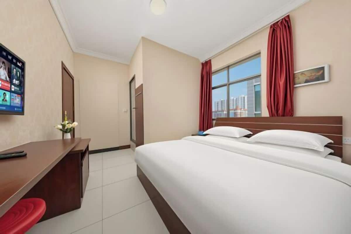 A hotel room with a bed, a desk with a chair, a TV on the wall, and a window with curtains.