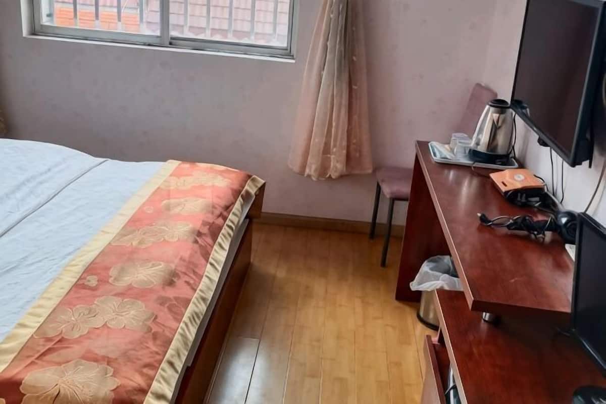 A bedroom with a bed, a desk with a computer, a chair, a TV, and a window with curtains.