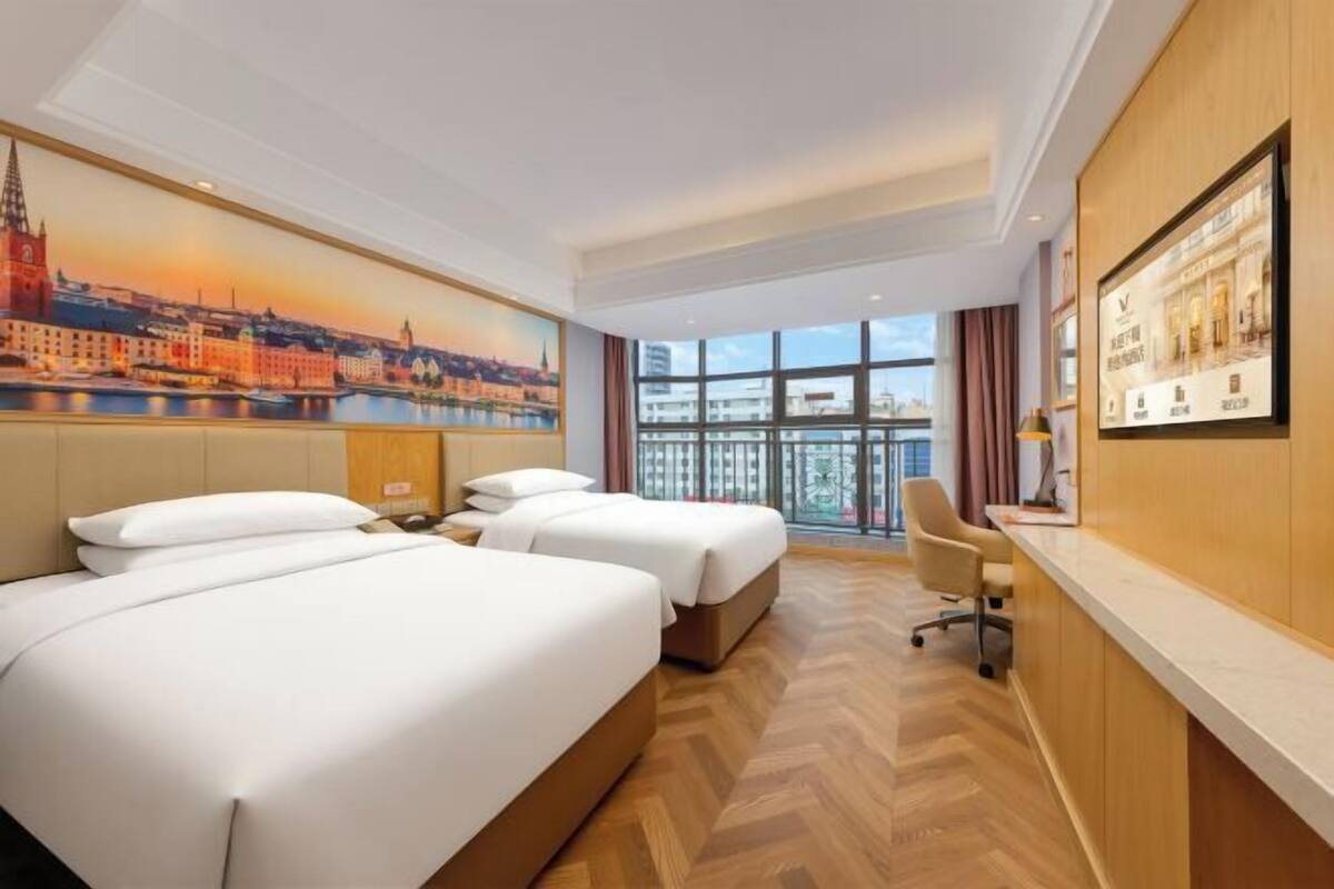 Ein Hotelzimmer mit zwei Betten, einem Schreibtisch und einem großen Fenster mit Blick auf die Stadt.