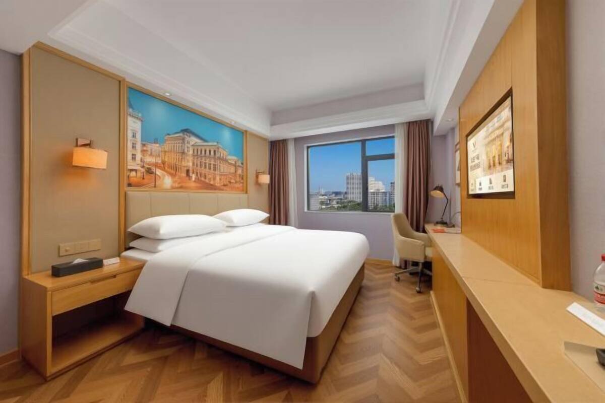 Ein Hotelzimmer mit Bett, Schreibtisch, Stuhl, Nachttisch, einer Wandlampe, einem Fenster mit Blick und einem Bild an der Wand.