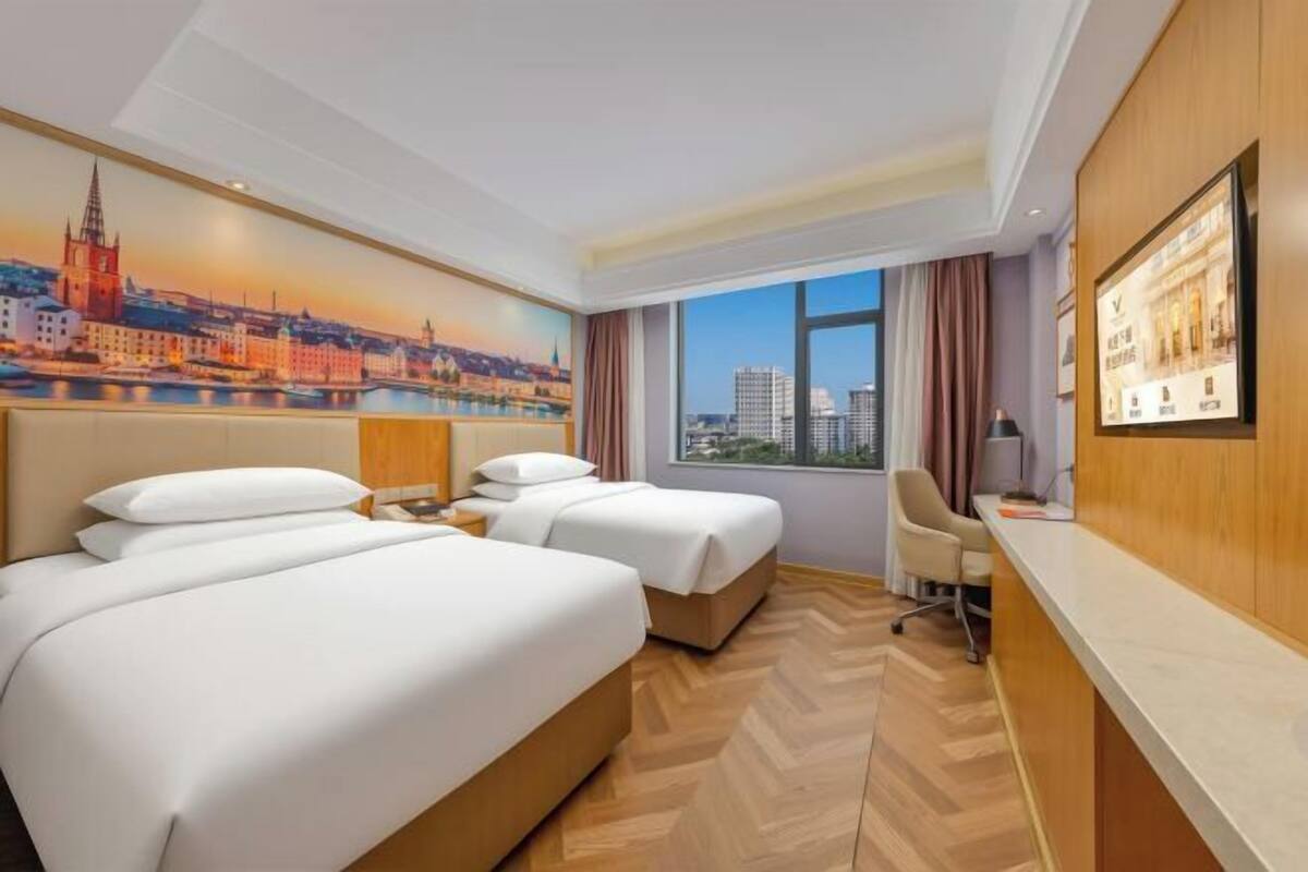 Ein Hotelzimmer mit zwei Betten, einem Schreibtisch, einem Stuhl und einem großen Fenster mit Blick auf die Stadt.