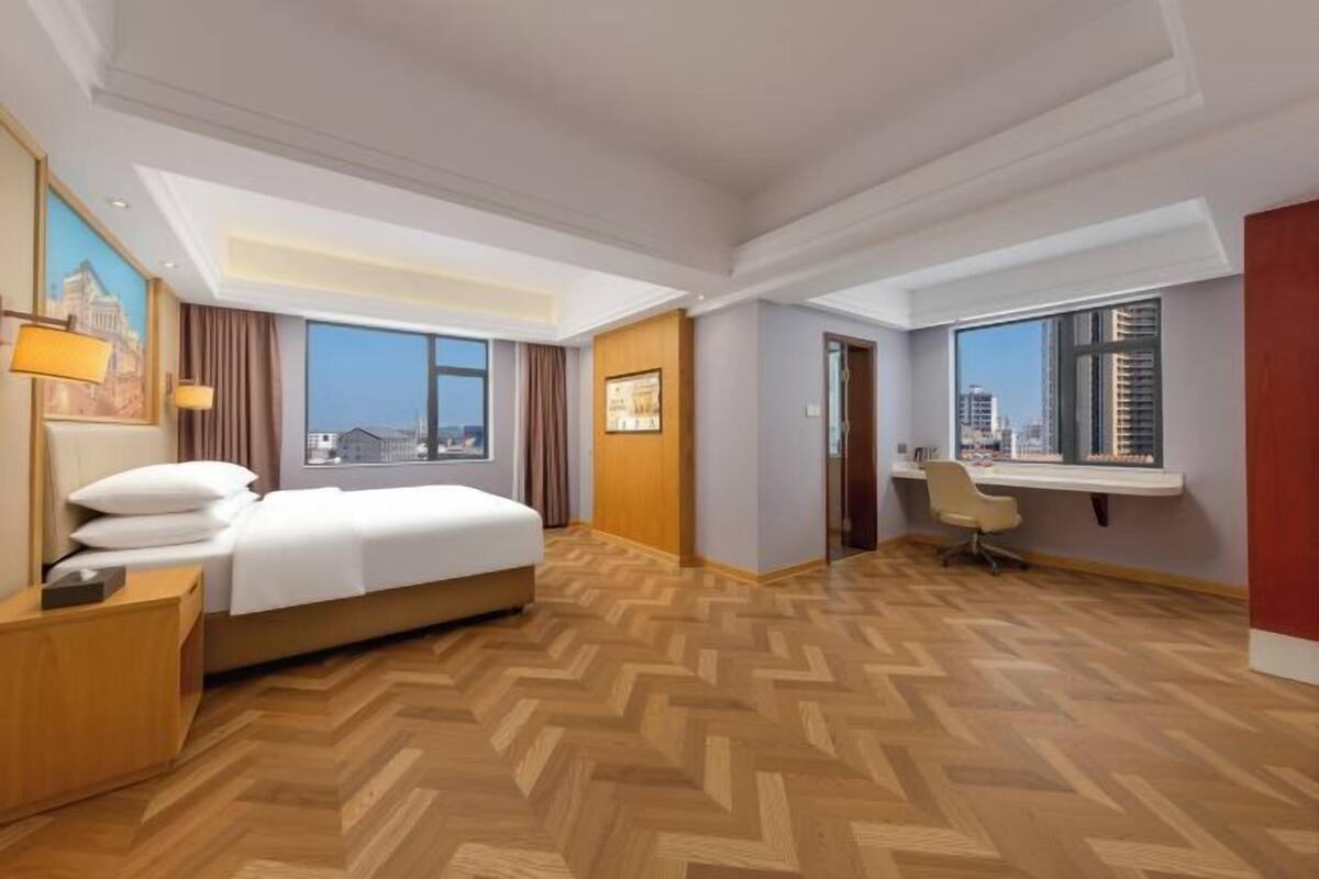 Ein Hotelzimmer mit Bett, Schreibtisch, Stuhl, großen Fenstern und Blick auf die Stadt.