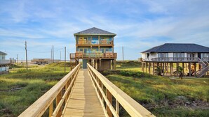 A wooden boardwalk leading to a two-story beach house with a balcony, a covered area with stairs, and a beach view. - Surfside Beachfront Paradise (Surfside Beach)