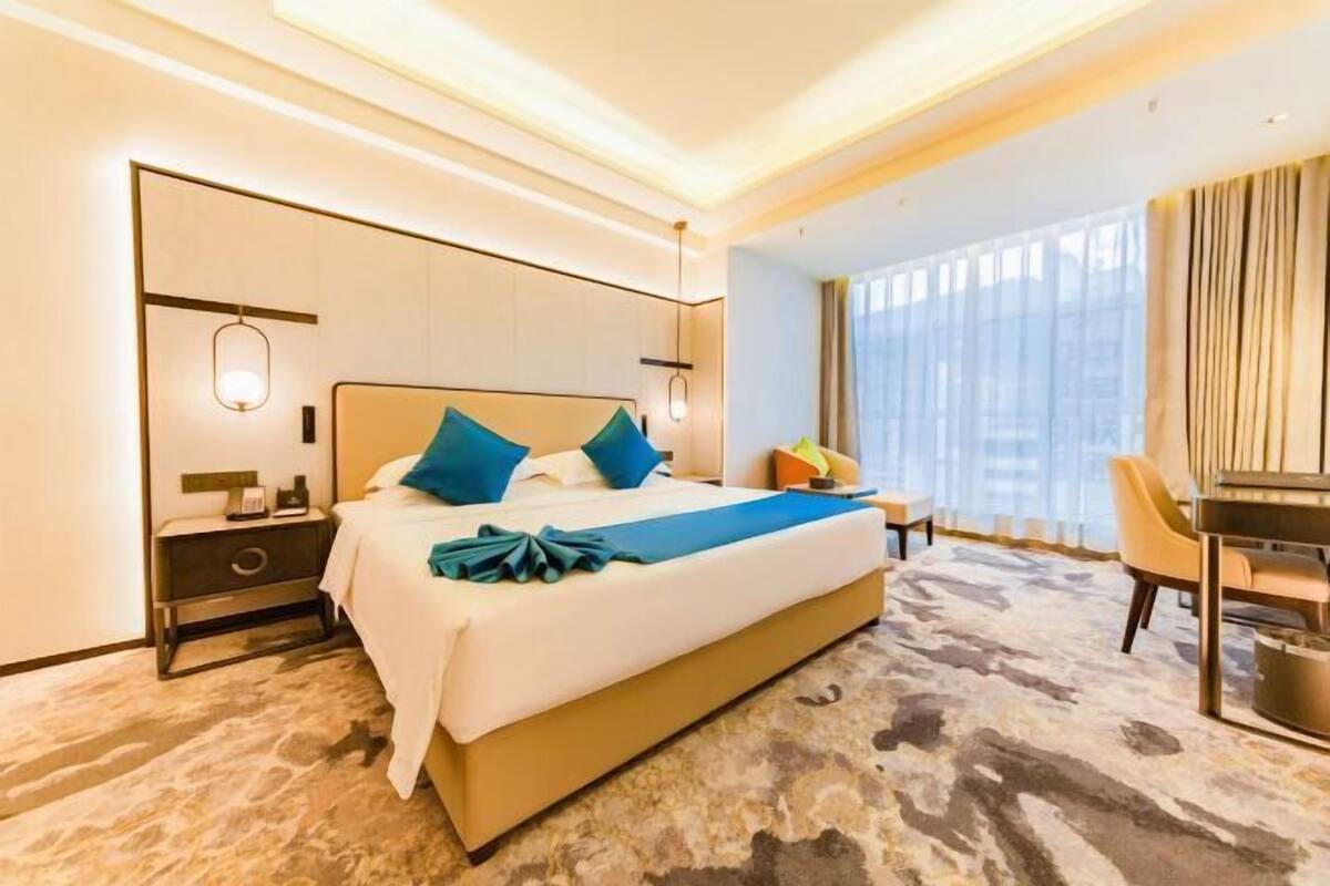 A hotel room with a large bed, bedside tables, a desk, and a view of the city through large windows.
