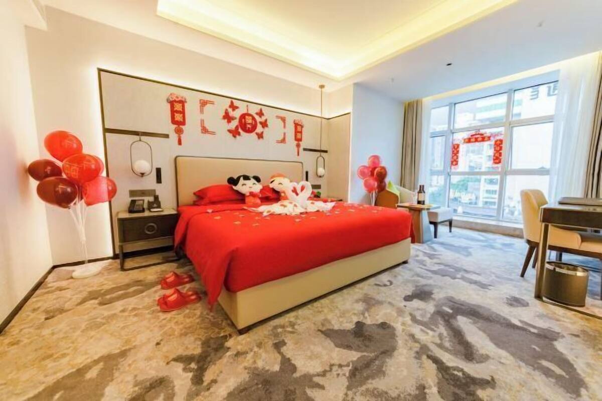 A hotel room with a large bed, a desk, a chair, a nightstand, and a window with curtains.