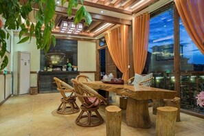 A dining area with a wooden table and chairs, a kitchen in the background, and a view through large windows.