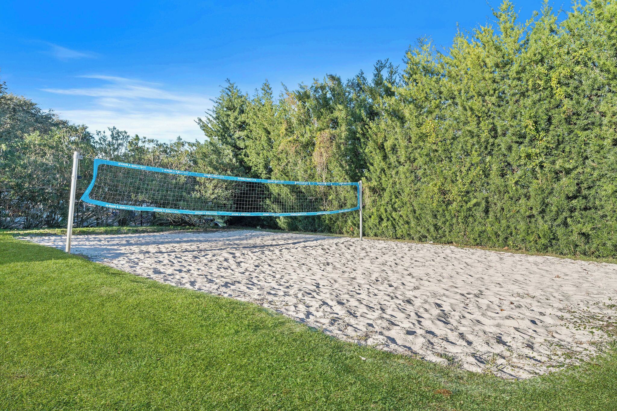 Un filet de volley-ball est installé sur un terrain de sable, entouré d’herbe verte et d’arbres.