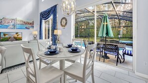 Un coin repas avec une table en verre, des chaises blanches et une vue sur une piscine, visible à travers de grandes fenêtres.