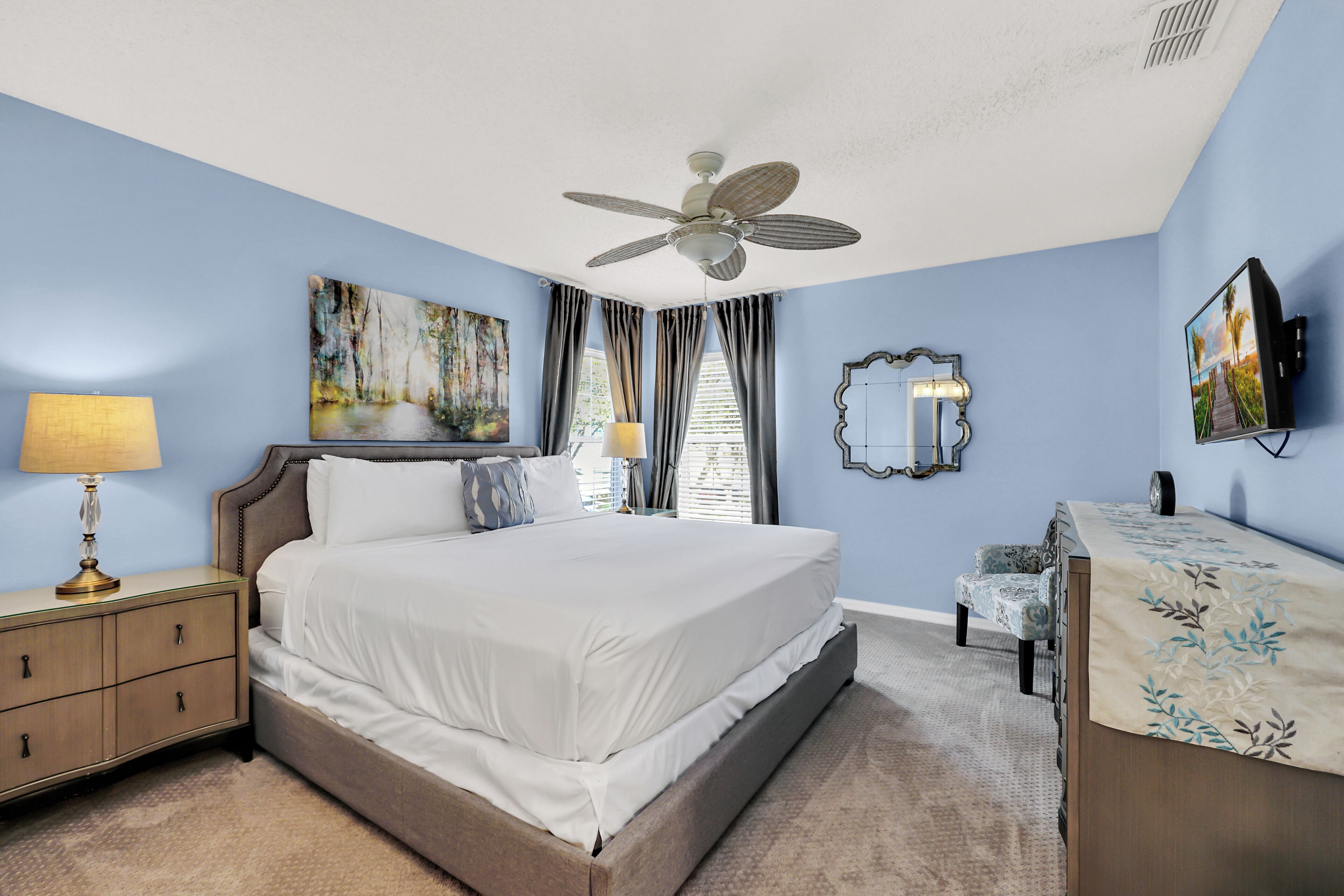 Une chambre à coucher avec un grand lit, une table de chevet avec une lampe, un ventilateur de plafond et des œuvres d’art accrochées aux murs.