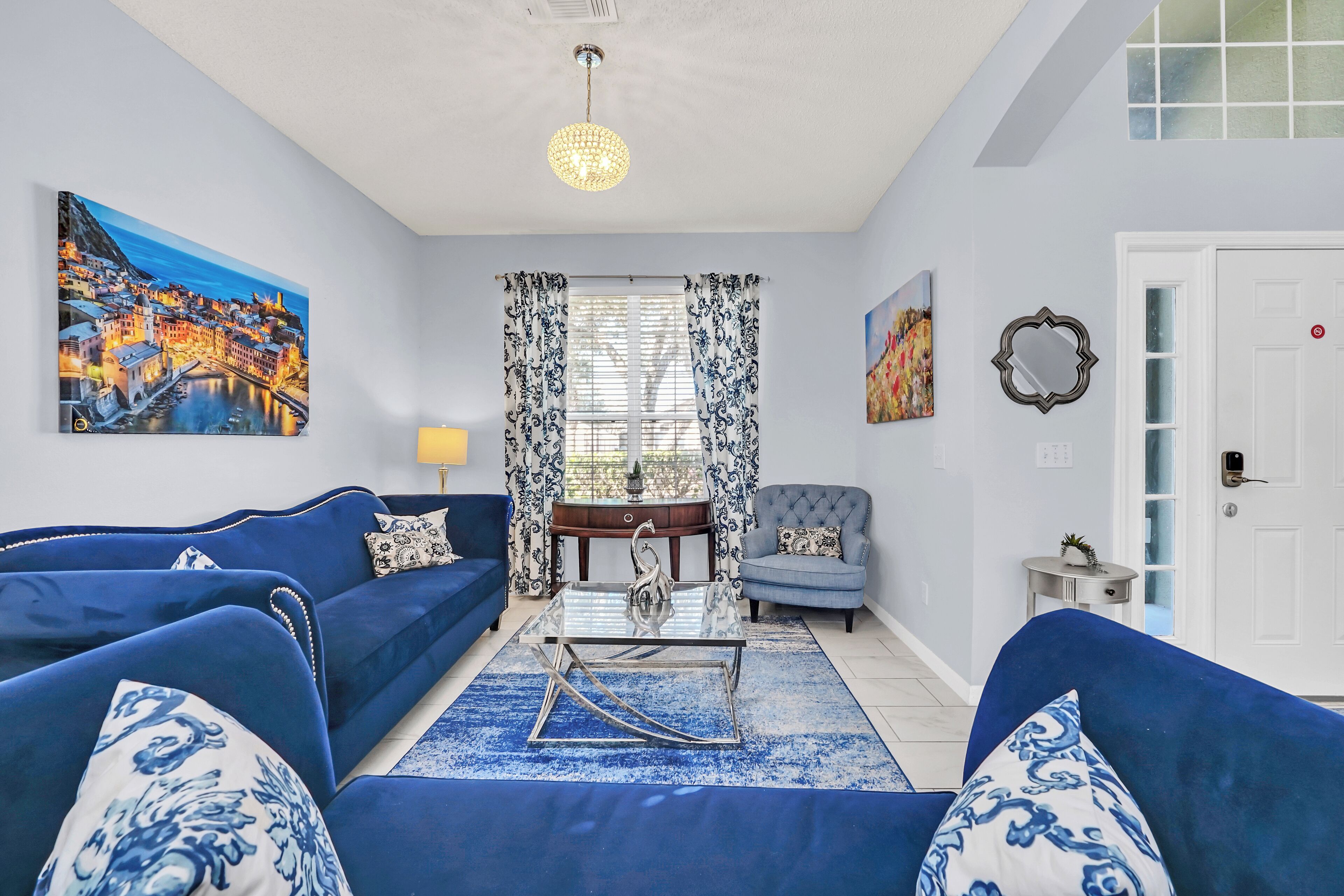 A living room with blue sofas, a glass coffee table, and a painting of a coastal town.