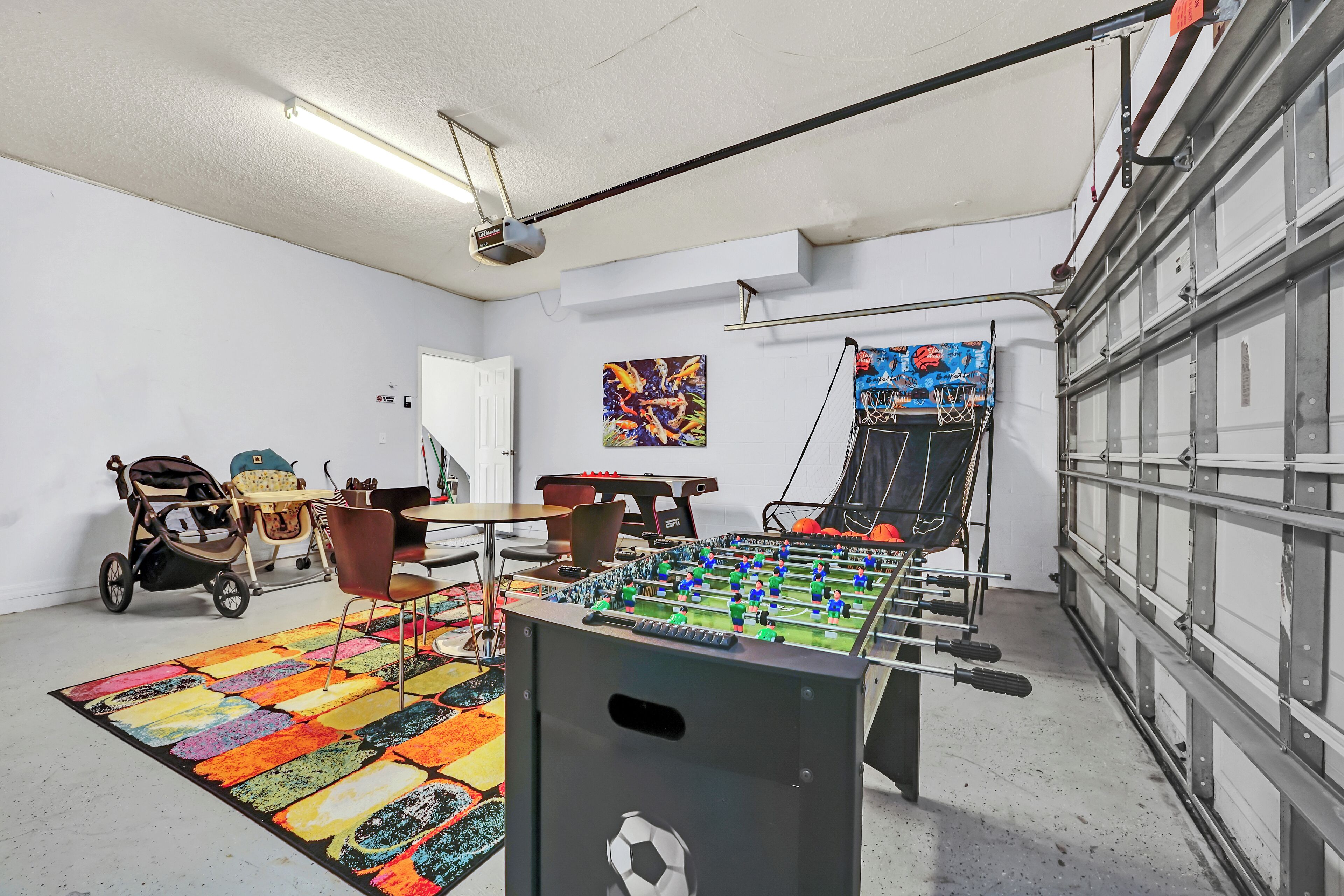 A room with a foosball table, a colorful rug, a stroller, and a basketball hoop.