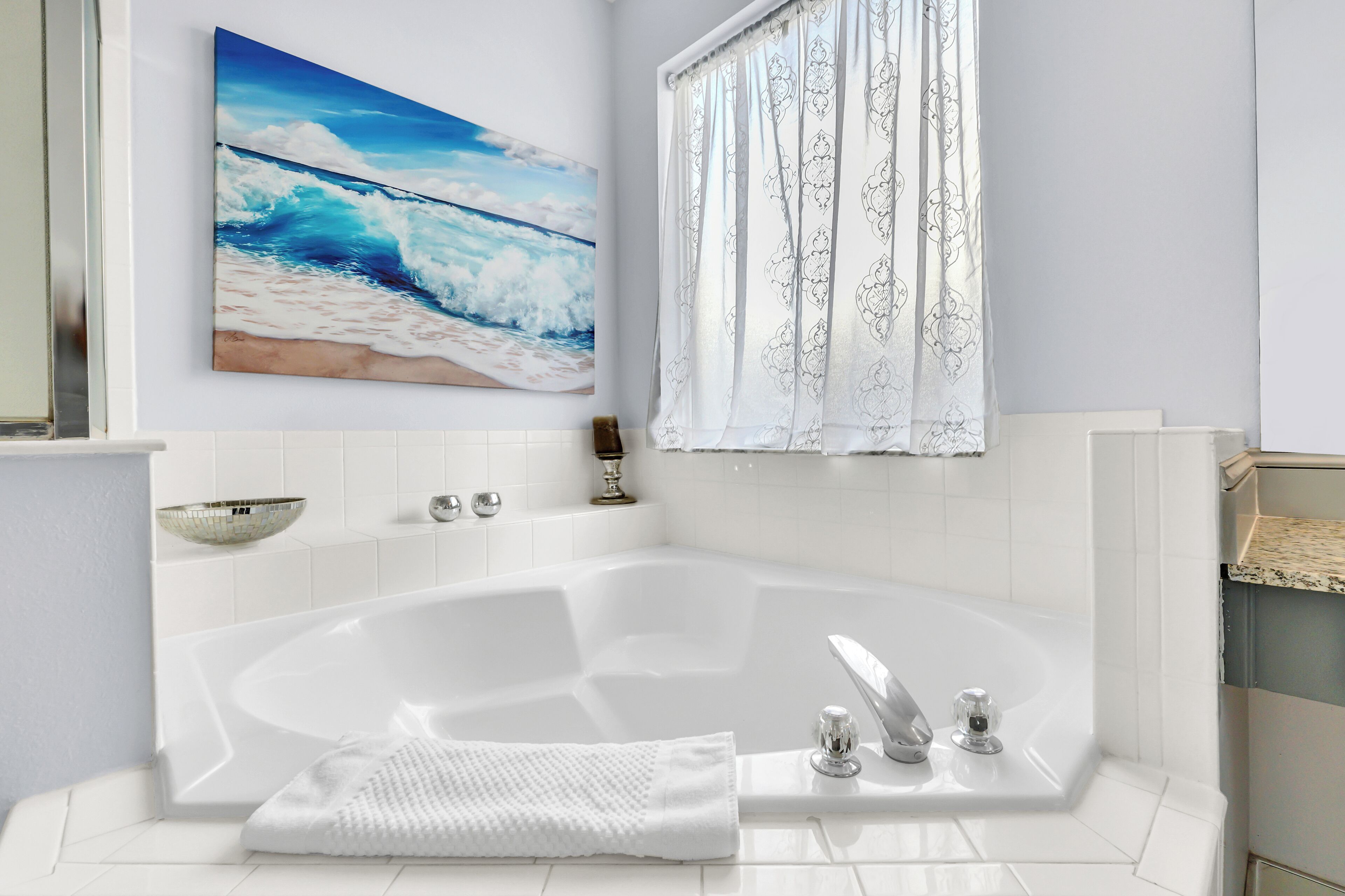 A modern bathroom with a large bathtub, a decorative wall painting, and a patterned window curtain.