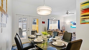A dining area with a glass table set for four, surrounded by black chairs. There is a ceiling fan, a potted plant, and a television on the wall. - 8953 Candy Palm Road (Kissimmee)