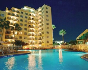 Un hôtel à plusieurs étages doté d’une piscine, de palmiers et d’une façade bien éclairée.