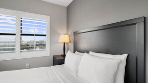 Habitación de hotel con cama, mesita de noche con lámpara, ventana con persianas y vistas a edificios.