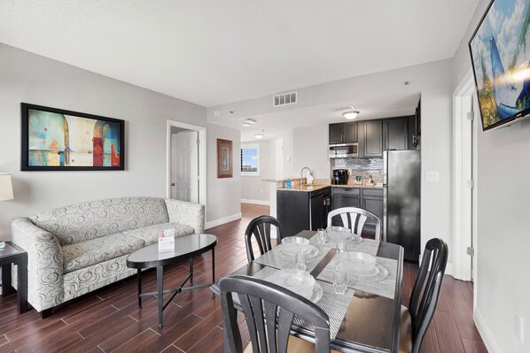 Un salon moderne avec un canapé, une table à manger et des chaises. On aperçoit une cuisine à l’arrière-plan, avec un réfrigérateur, une cuisinière et des placards.
