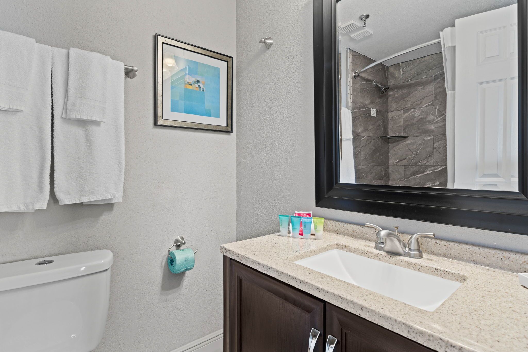 A bathroom with a white sink, dark wooden cabinets, a framed picture on the wall, and a shower area visible through the mirror.