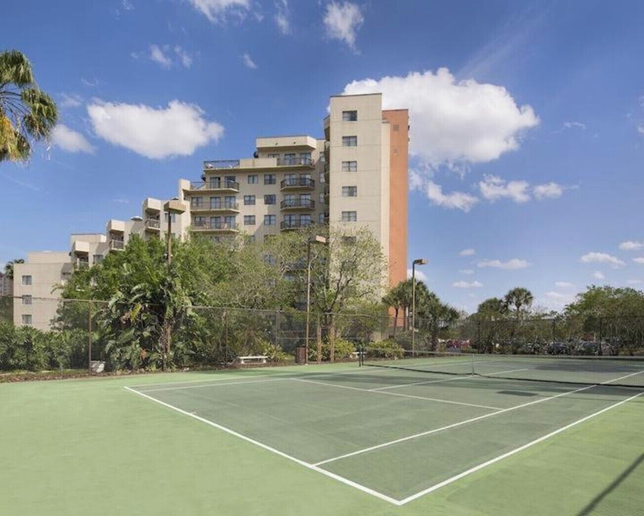 A tennis court with a net, surrounded by greenery and a multi-story building with balconies.