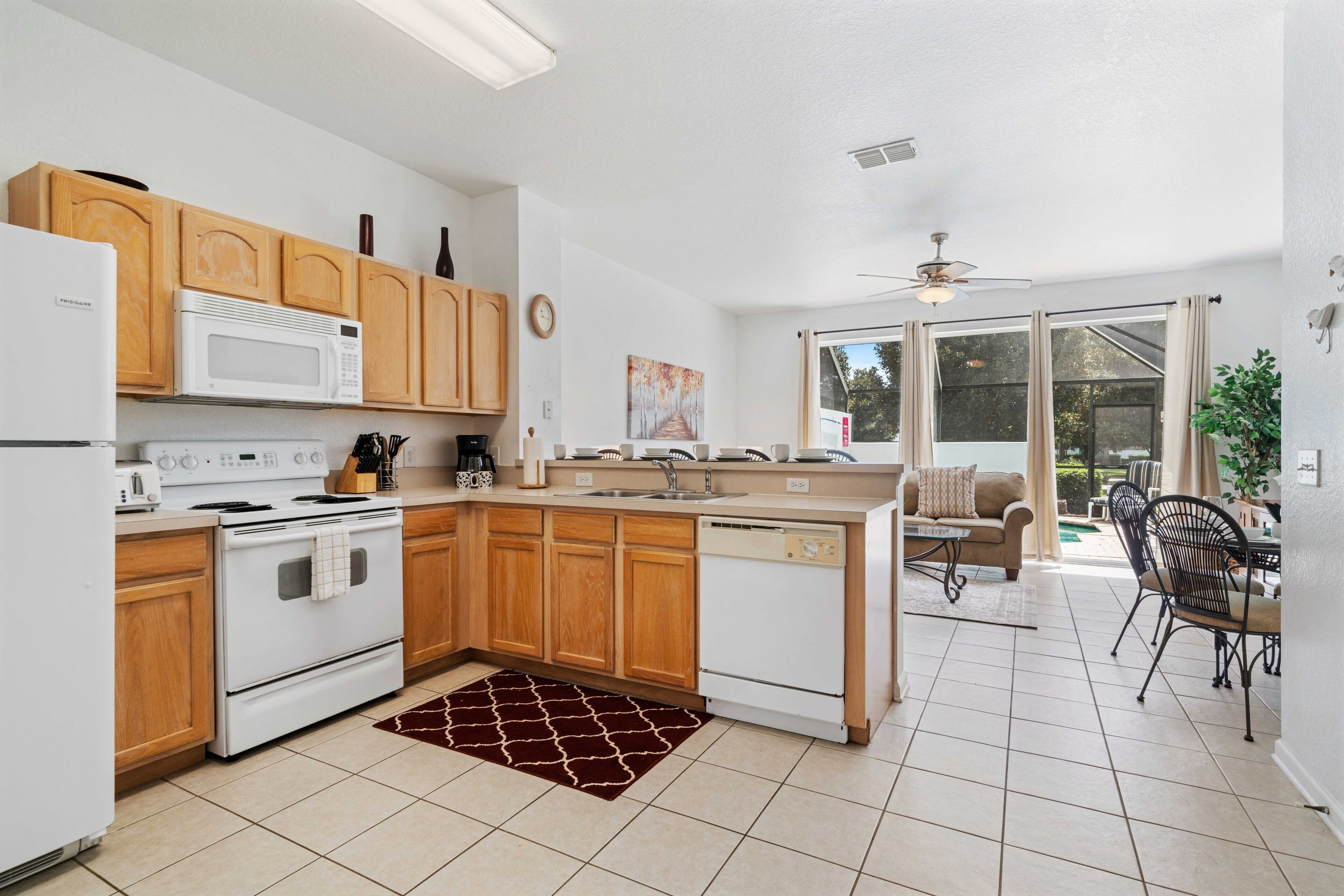 A modern kitchen with wooden cabinets, a white stove, and a dishwasher. There is a dining area with a table and chairs, and a view of the outdoors through large windows.