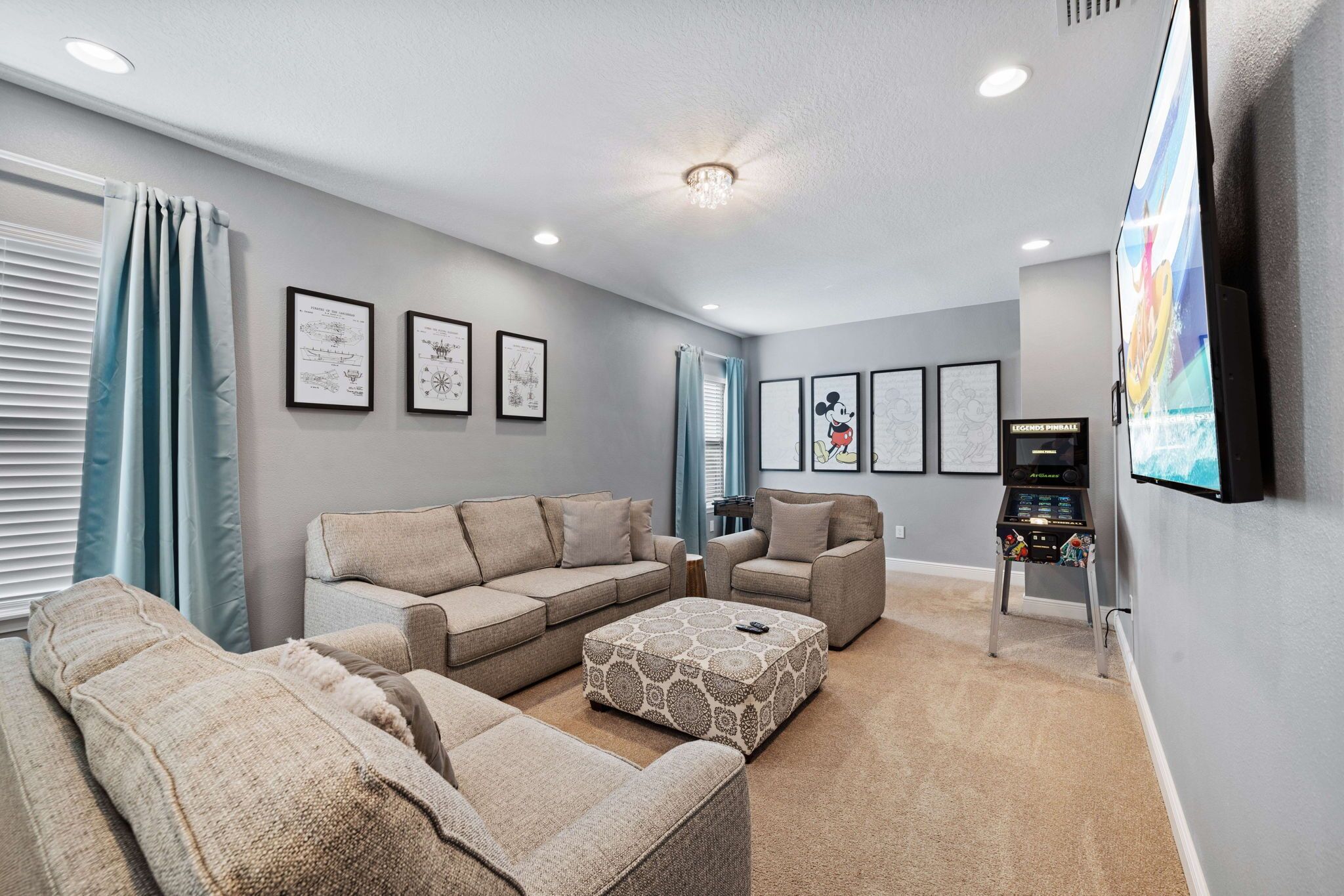 A living room with a beige sectional sofa, a patterned ottoman, and a flat-screen TV mounted on the wall.