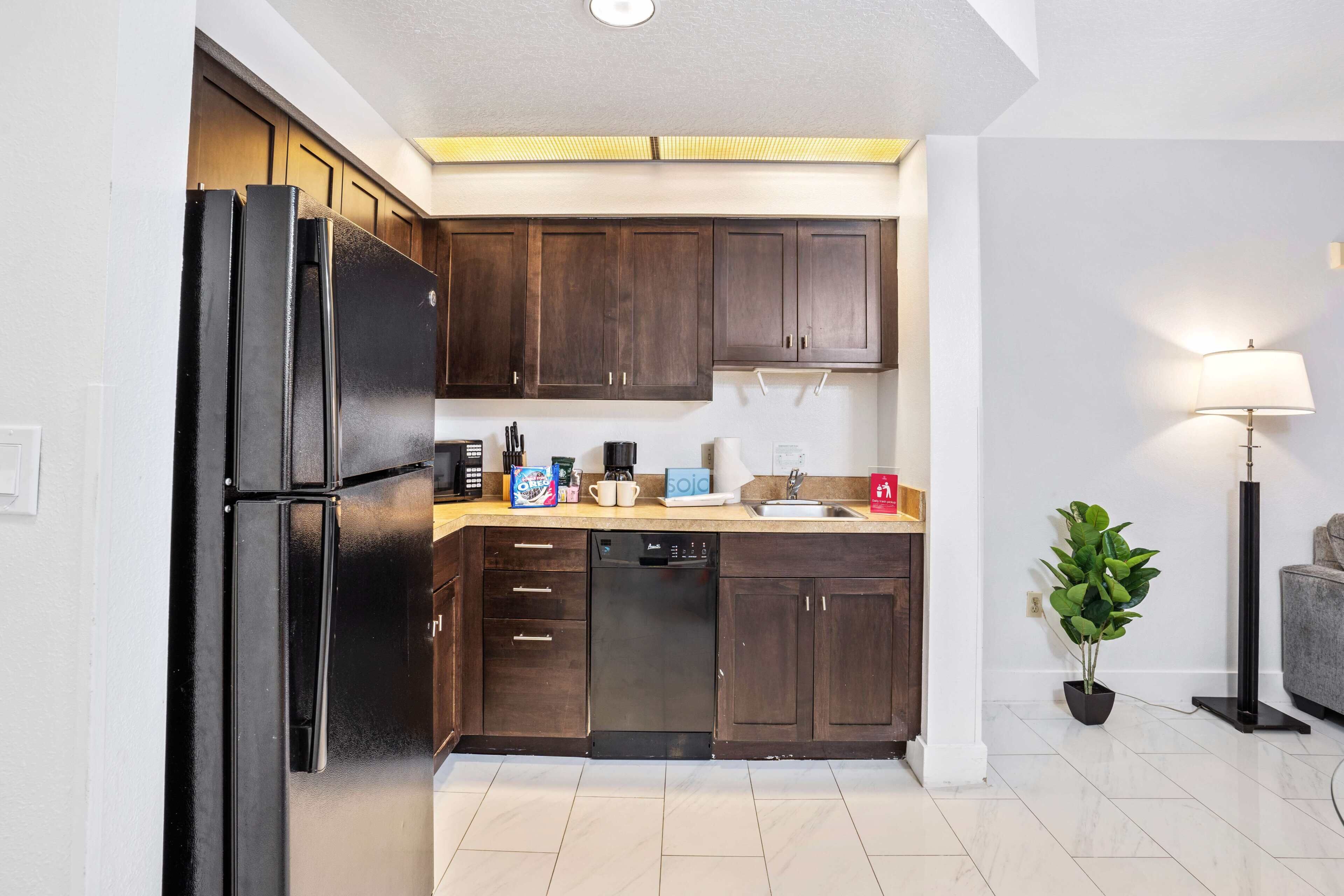 Una cocina moderna con armarios de madera oscura, un refrigerador negro y un lavavajillas. Hay un lavamanos con grifo, una encimera con varios objetos y una planta en maceta.