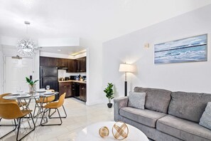 Ein modernes Wohnzimmer mit einem grauen Sofa, einem Esstisch mit Stühlen, einem Couchtisch, einer Stehlampe und einem Gemälde mit Strandmotiv.