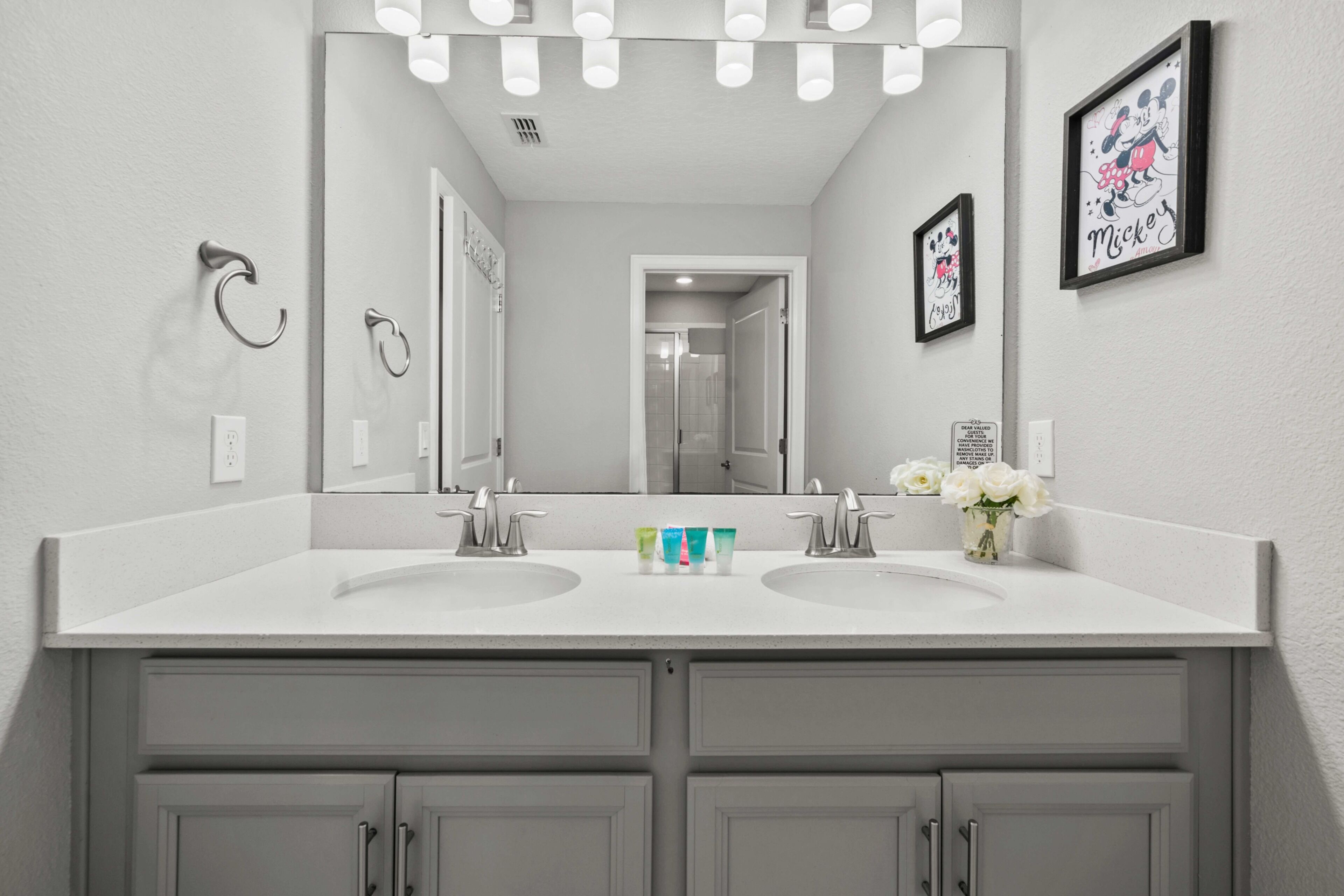 Une salle de bain avec deux lavabos, un grand miroir et un tableau encadré représentant Mickey Mouse accroché au mur.