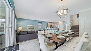 A dining area with a wooden table set for six, white upholstered chairs, and a modern chandelier. - 9046 Stinger Drive (Davenport)