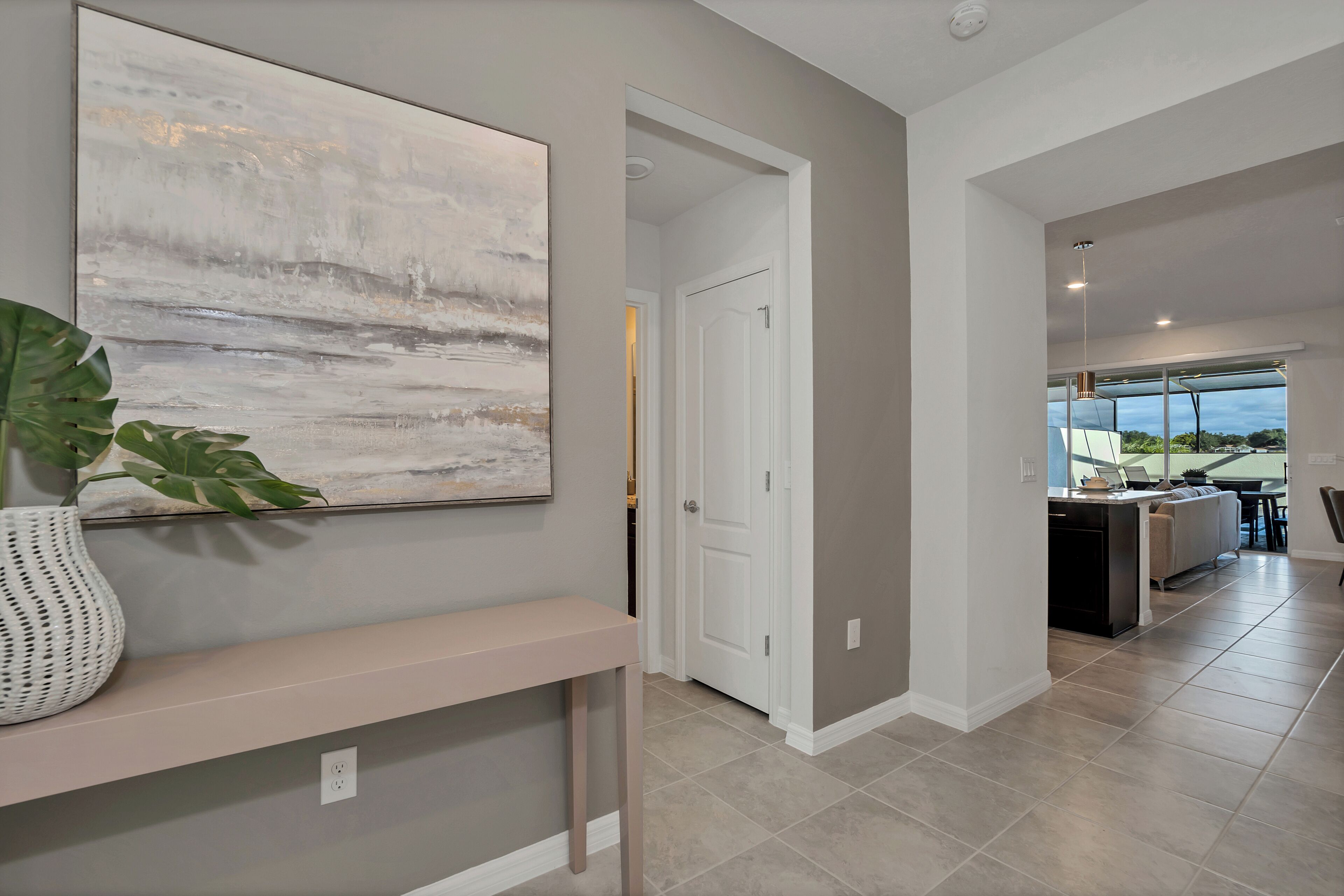 A modern interior with a neutral color palette, a large abstract painting, a potted plant, and a view into a dining area.