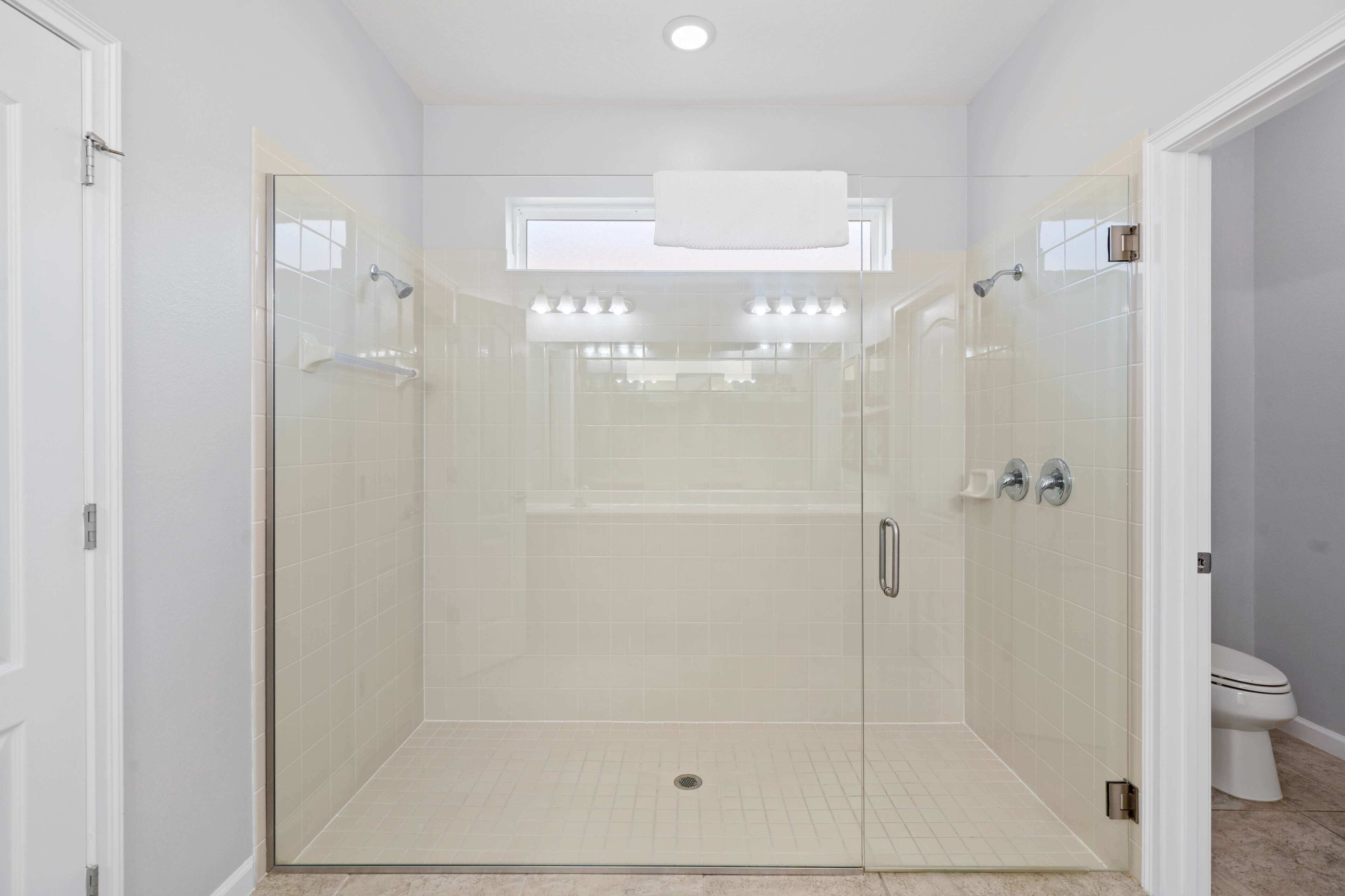 A glass-enclosed shower stall with a white towel hanging above the door, a tiled floor, and a visible toilet in the adjacent room.