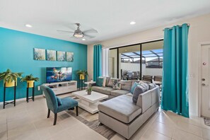 A modern living room with a teal accent wall, a ceiling fan, a flat-screen TV, a sofa, a coffee table, and a dining area visible through a glass door. - 8918 Sydney Avenue (Kissimmee)