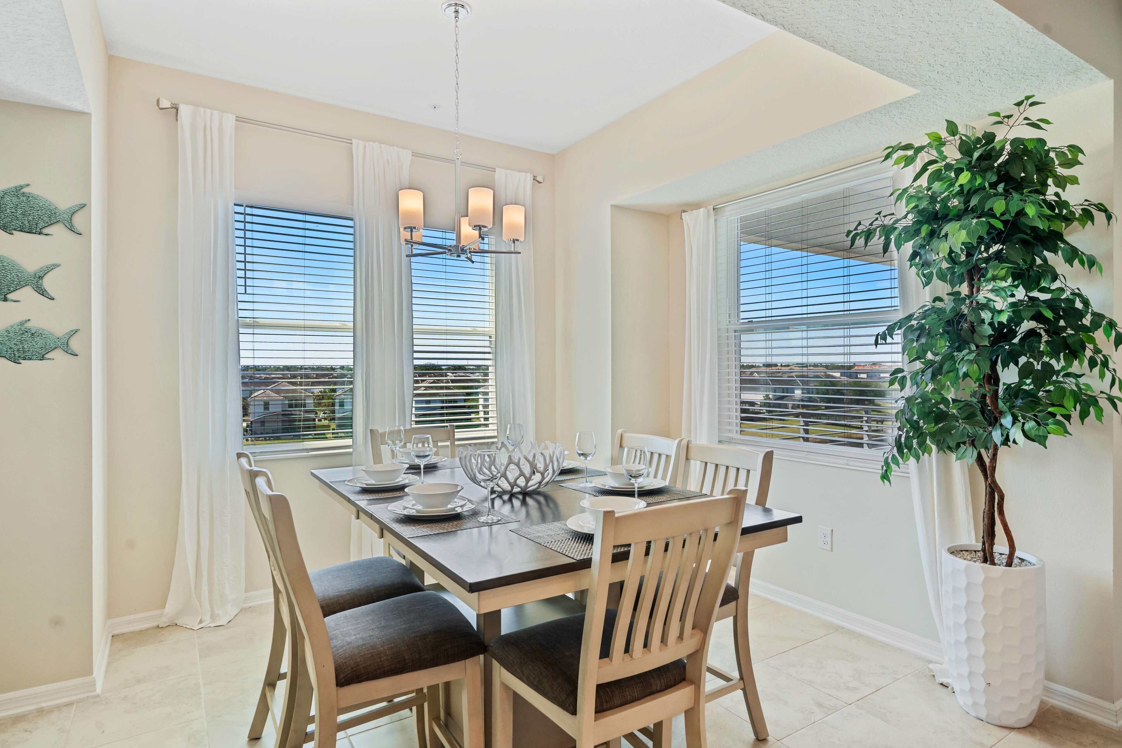 A dining area with a table set for four, large windows with blinds, a chandelier, and a potted plant.