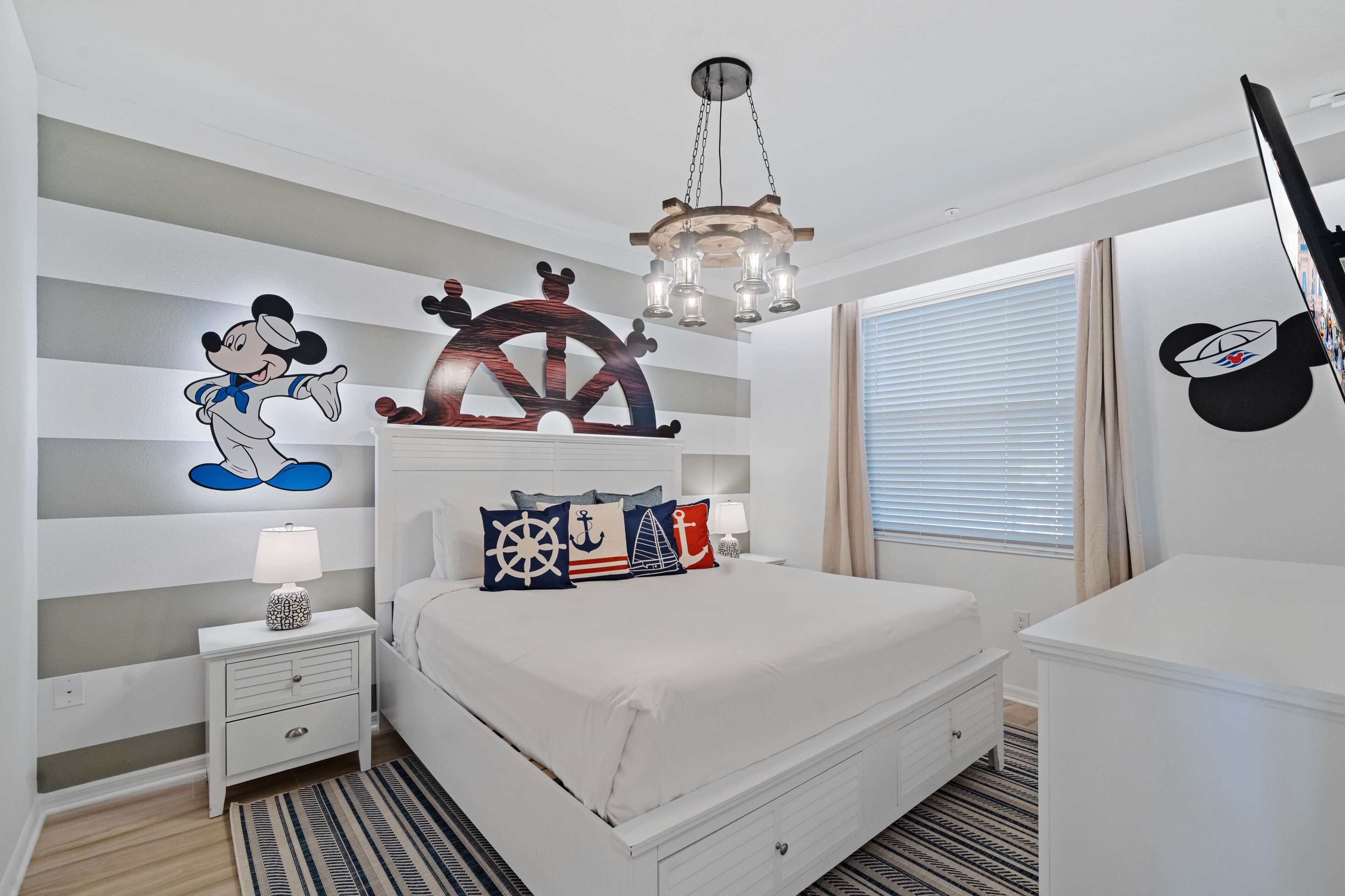 Une chambre avec un lit, une table de chevet, une lampe, un autocollant mural représentant Mickey Mouse, un élément de décoration en forme de roue de navire et un téléviseur.