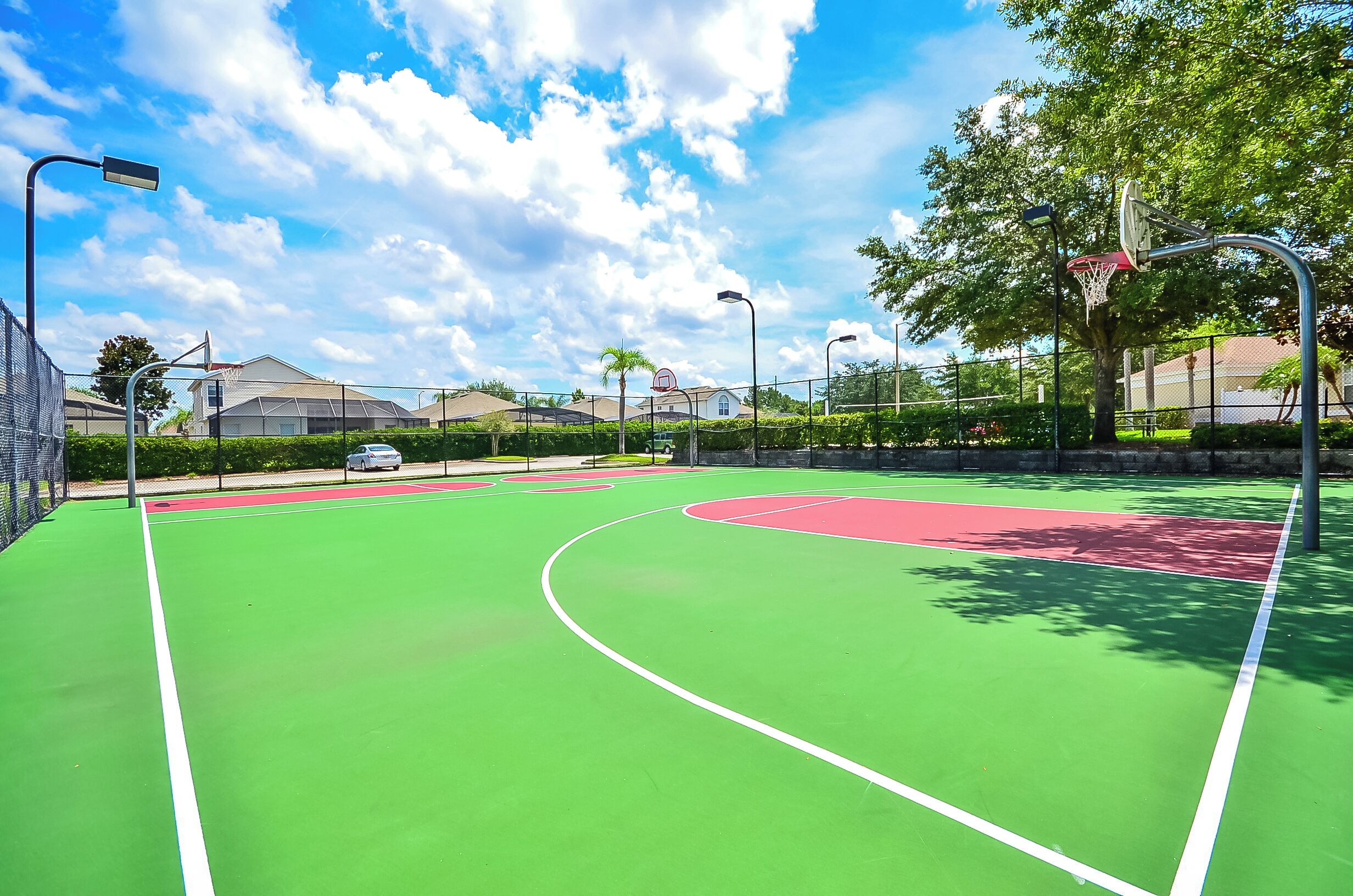 An outdoor basketball court with green and red markings, surrounded by trees and a fence.