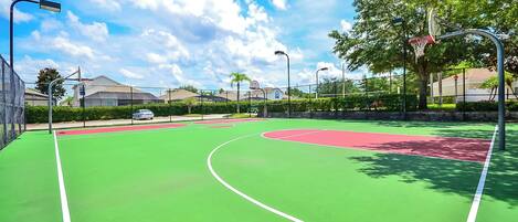 An outdoor basketball court with green and red markings, surrounded by trees and a fence.