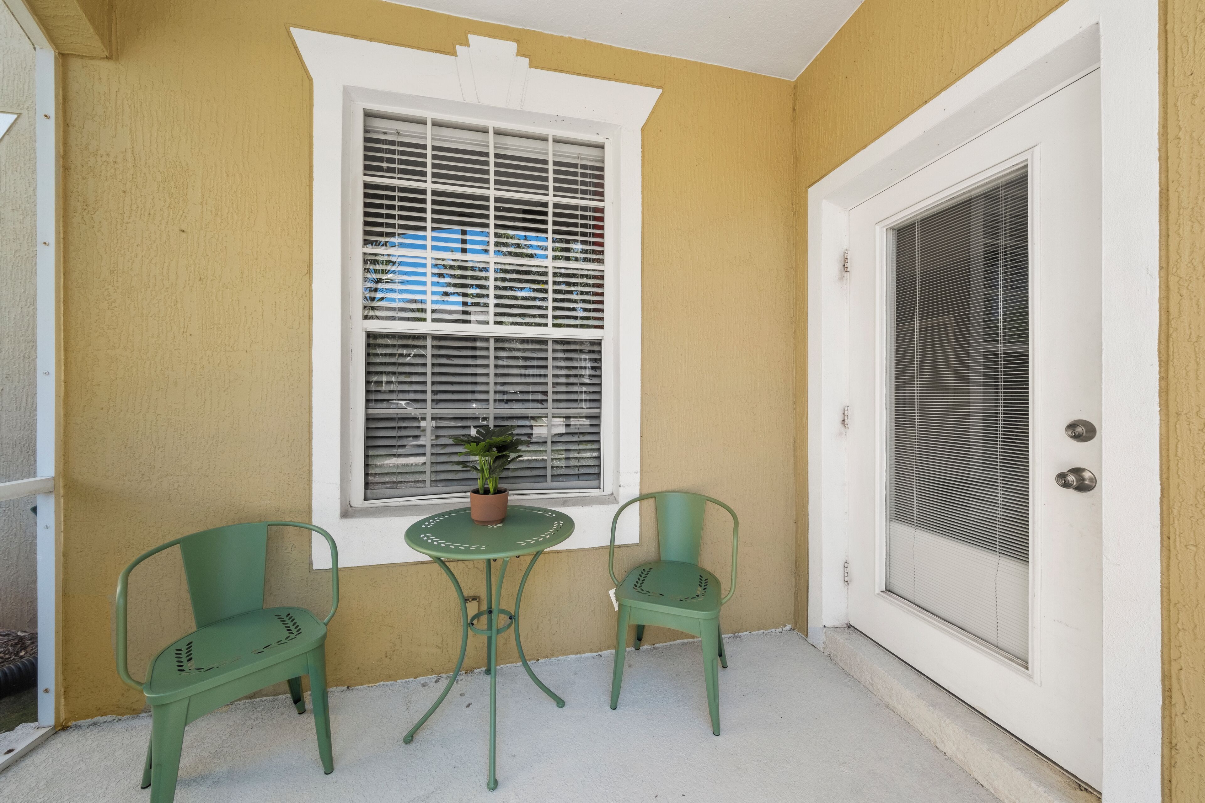A small patio with a white door, a window with blinds, and two green chairs.