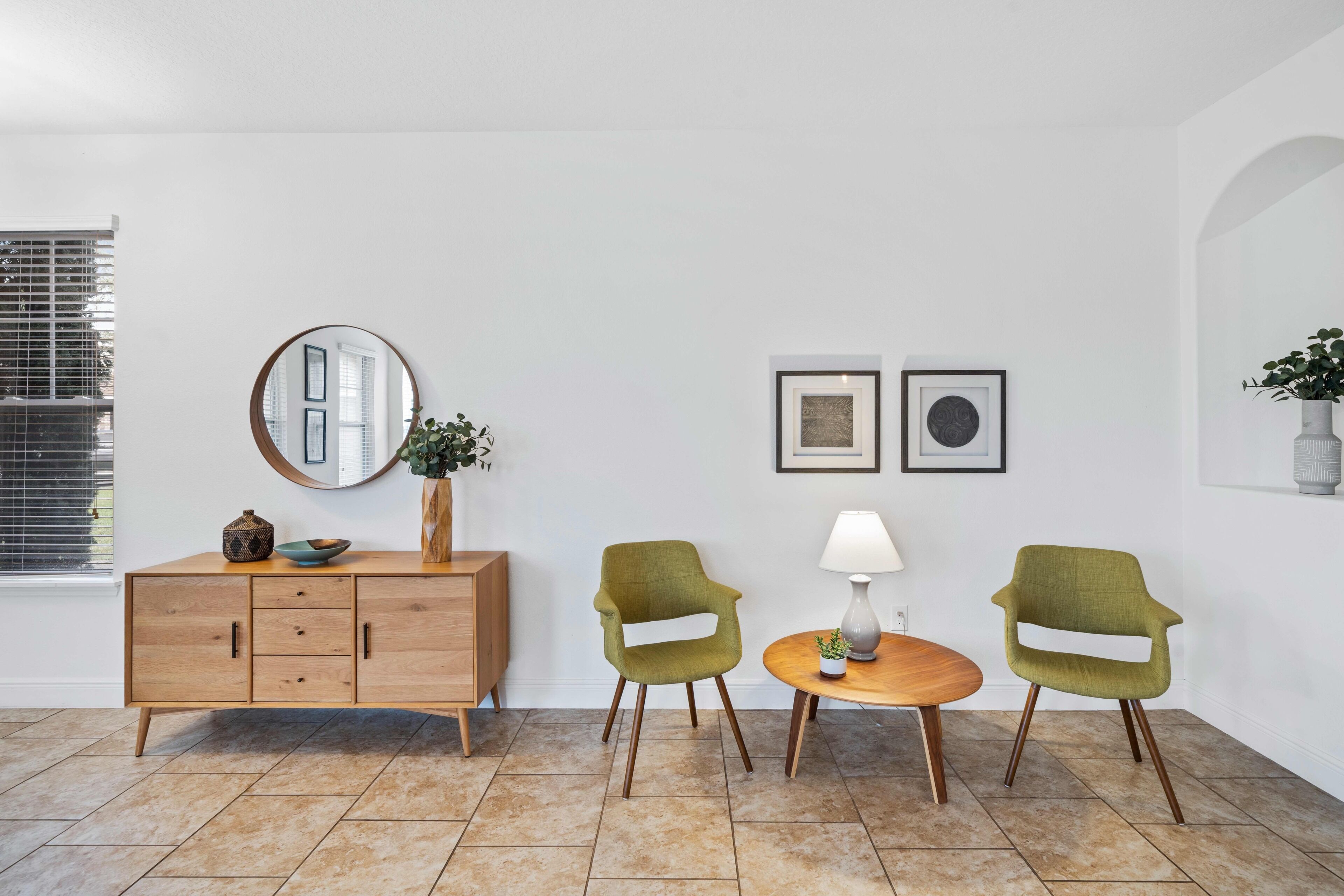 A room with a wooden cabinet, two green chairs, a round table with a lamp, and a mirror on the wall.