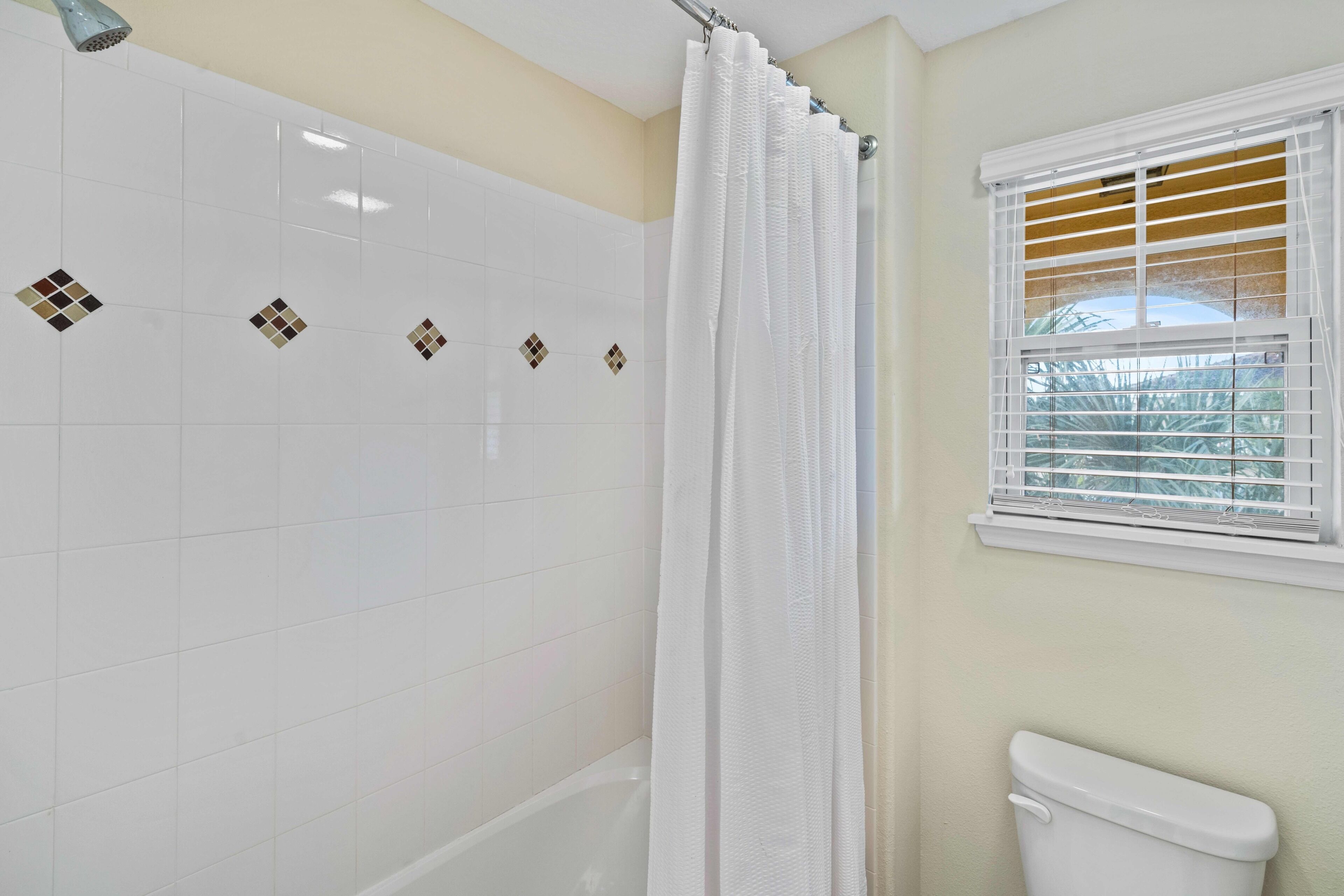 A bathroom with white tiles, a bathtub, a toilet, and a window with blinds.