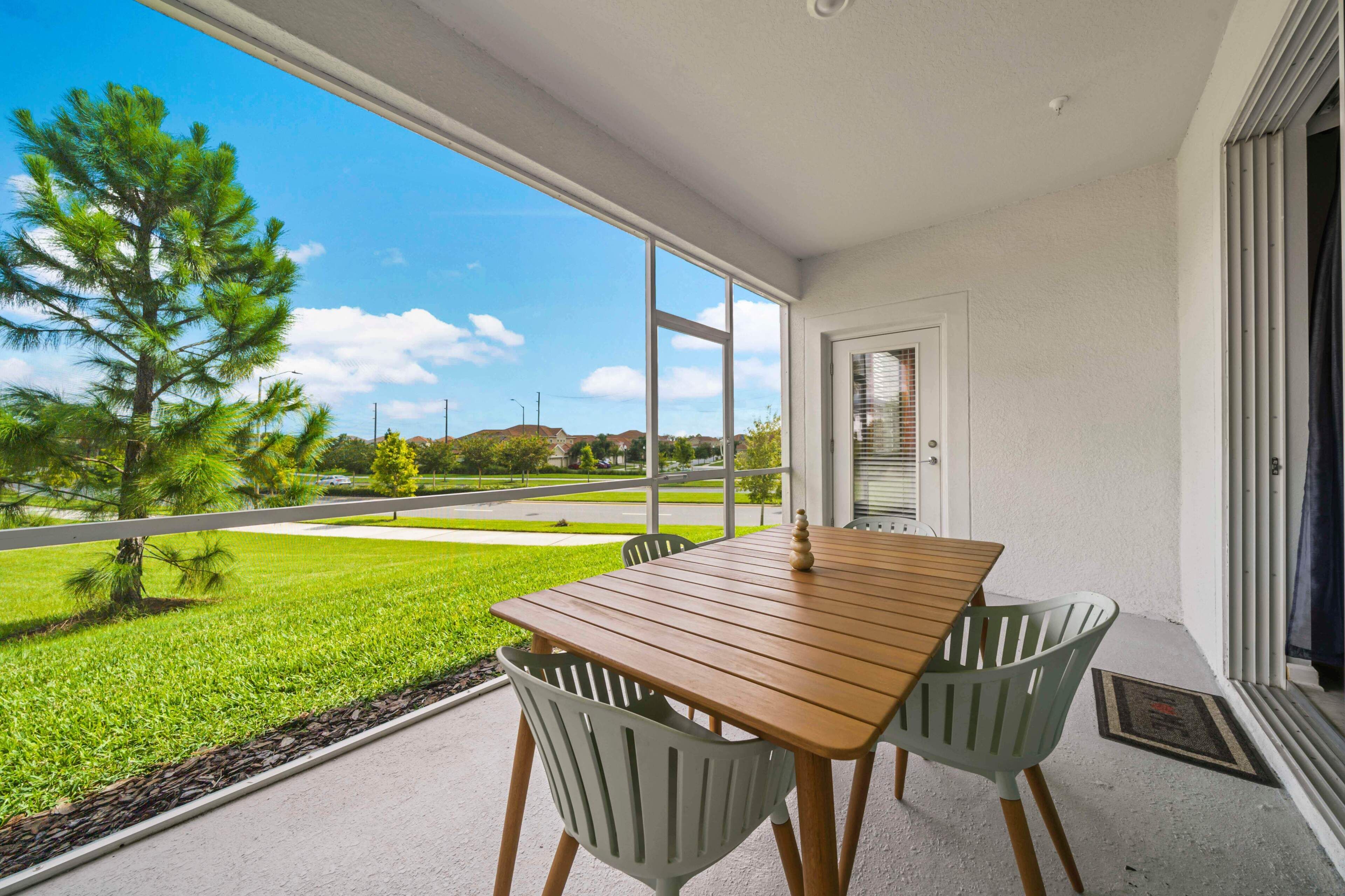 Varanda com mesa de madeira e duas cadeiras, com vista para um gramado verde e uma estrada.