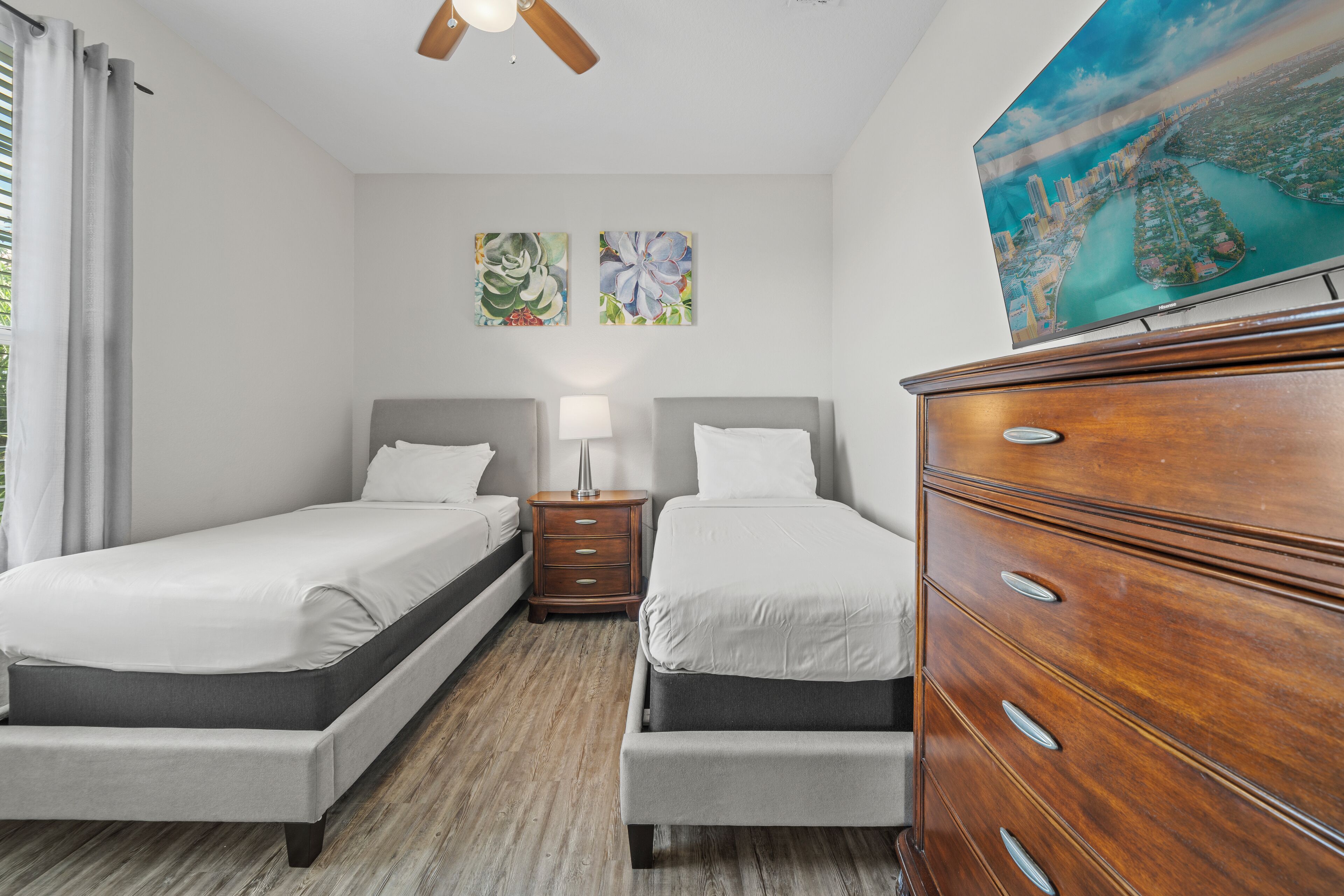 Une chambre d’hôtel avec deux lits simples, une commode en bois, une table de chevet avec une lampe et un ventilateur de plafond.