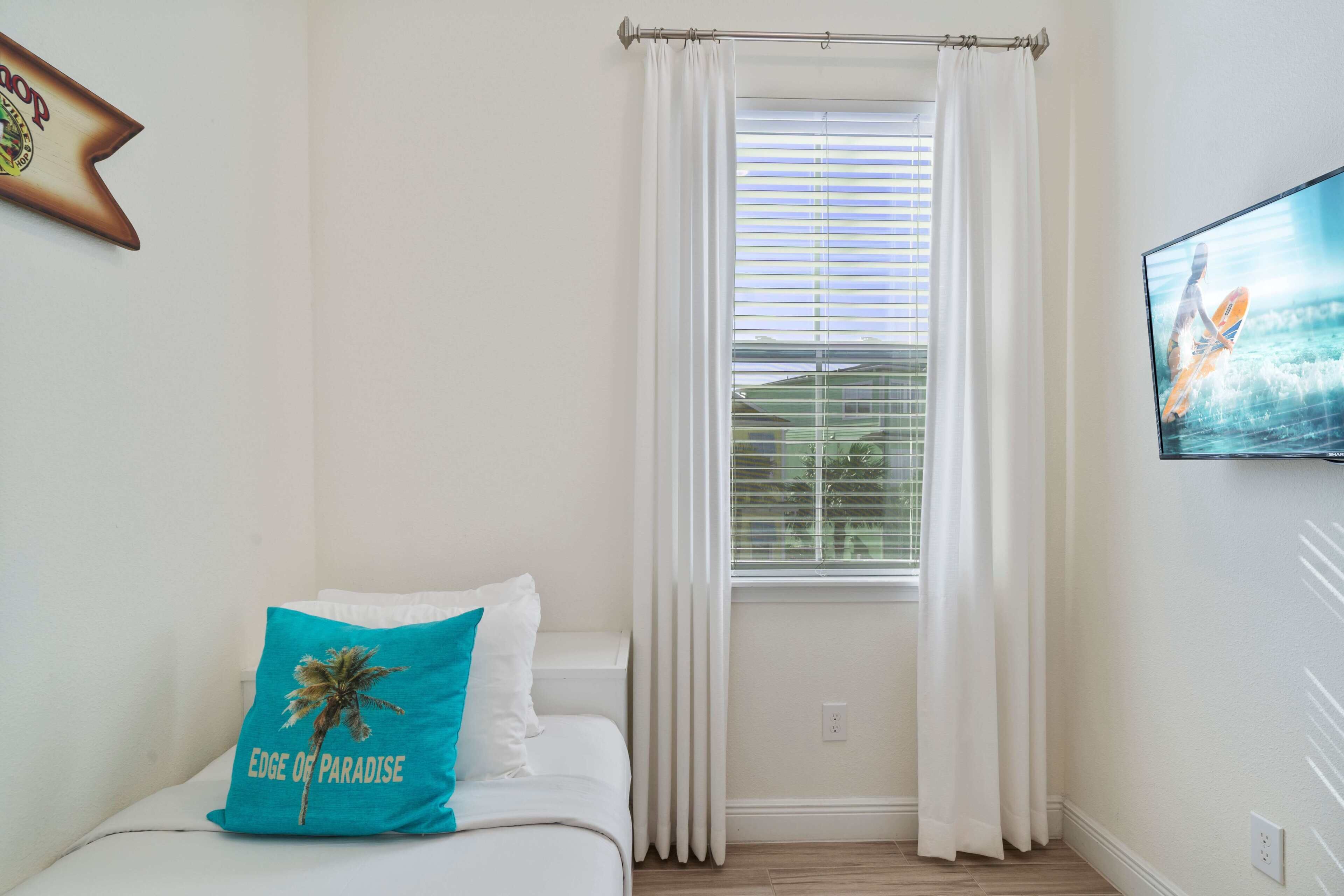 A bedroom with a bed, a window with white curtains, a mounted TV, and a wall-mounted picture frame.