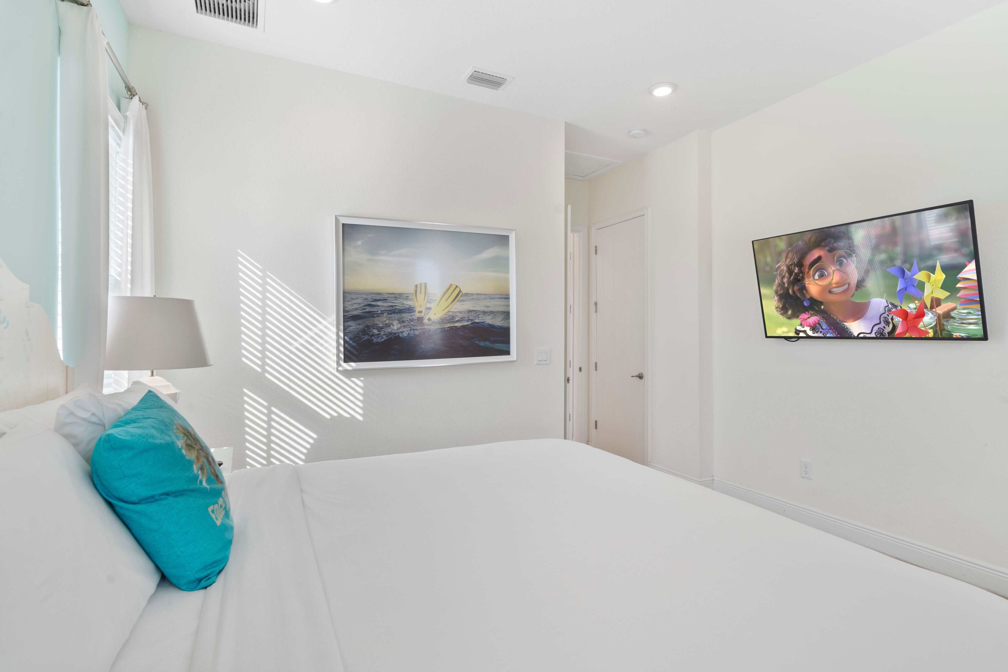 A bedroom with a bed, a teal pillow, a painting of a sunset, and a framed picture on the wall.