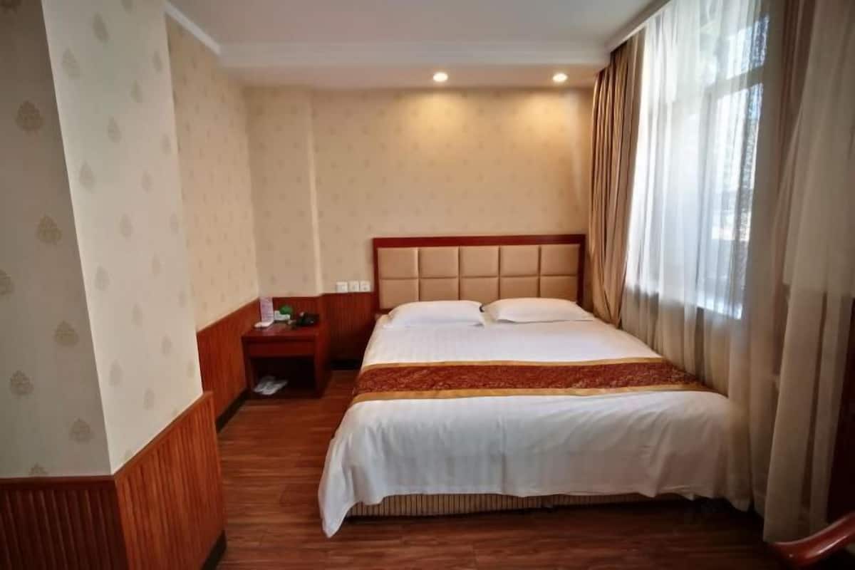 A hotel room with a large bed, a wooden nightstand, a wall-mounted telephone, and a window with curtains.