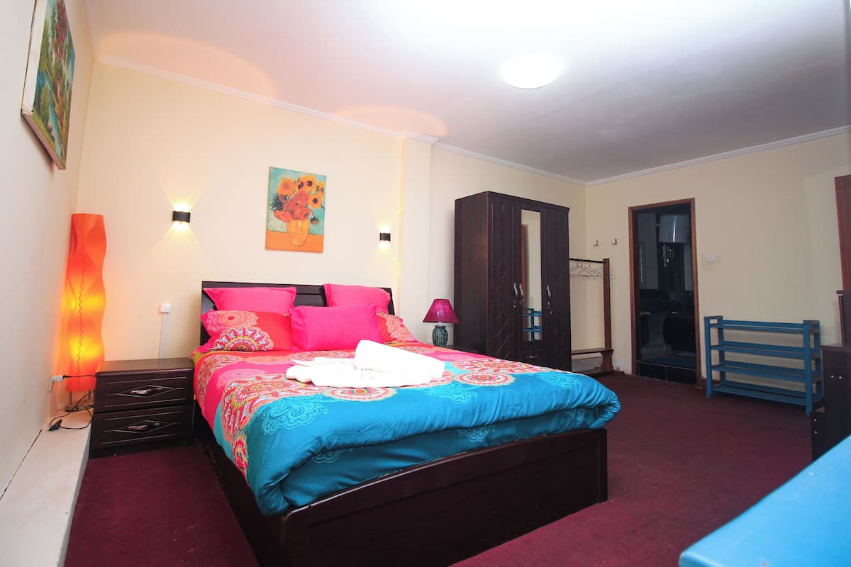 Una cama bien hecha con un colorido edredón azul y rosa, un armario de madera y una mesita de noche con una lámpara.