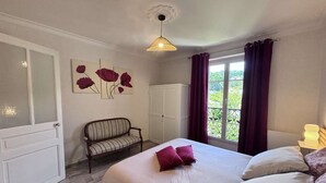 A bedroom with a bed, a bench, a wardrobe, a window with curtains, and a wall with floral artwork.
